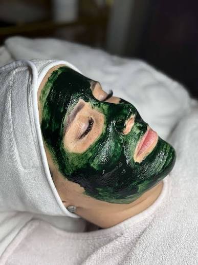 A dark green KRX Green Sea Peel spicule mask applied across a client's face during treatment, with a white towel headwrap.